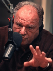 Man speaking into a microphone, gesturing hand.